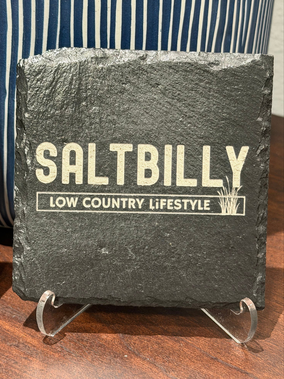 Sealed slate coaster with a dark finish, laser engraved with the Saltbilly Low Country Lifestyle logo on a clear display stand