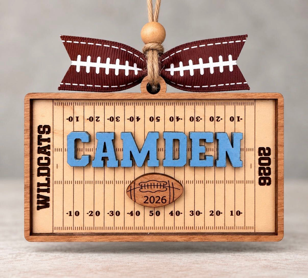 Rectangular wooden football field ornament with yard lines, a name in blue, team name, and year, tied with a football ribbon