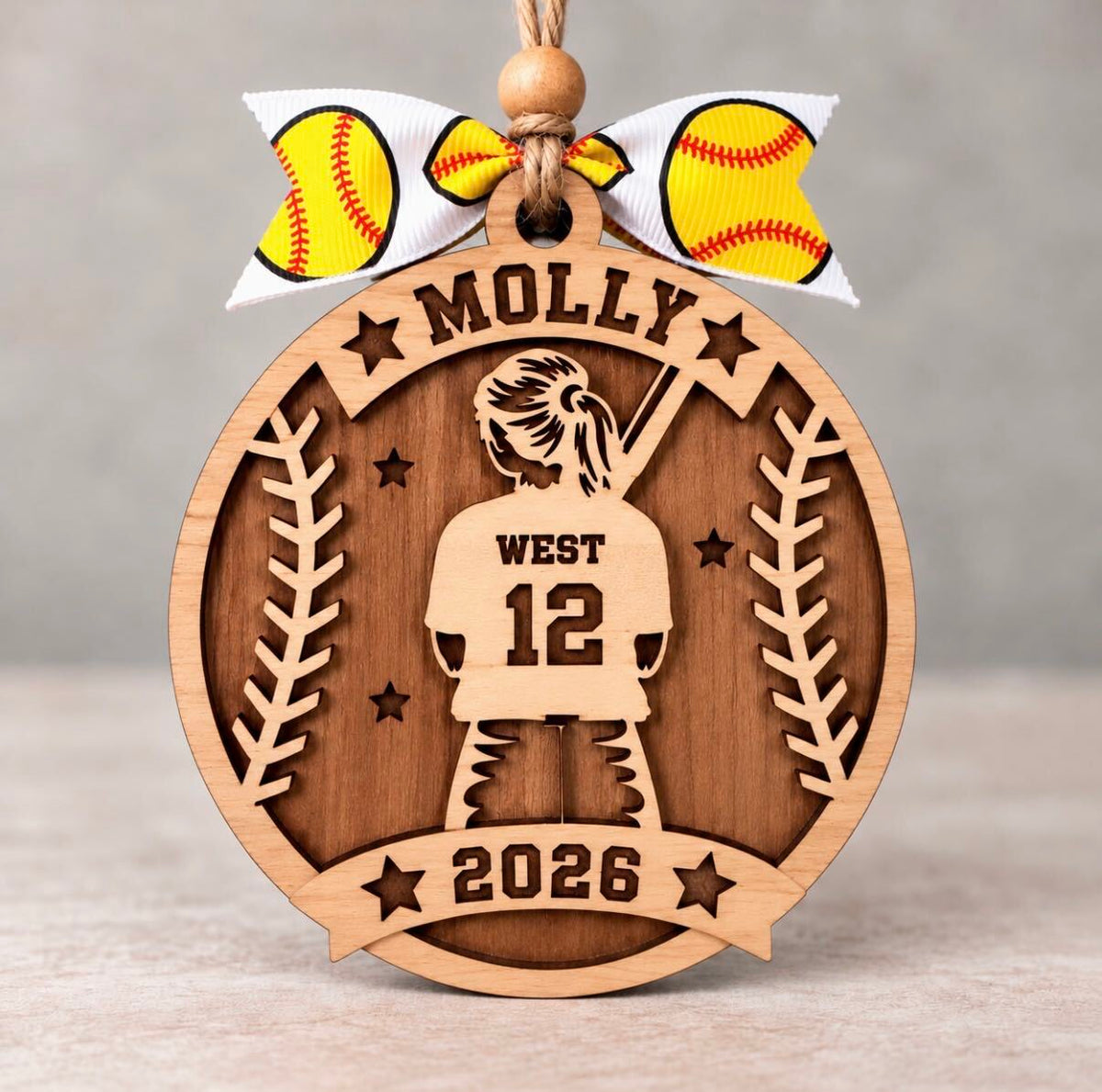 Round layered wood softball ornament with a ponytailed player holding a bat, personalized with Molly, West, number 12, and 2026, tied with a yellow softball ribbon