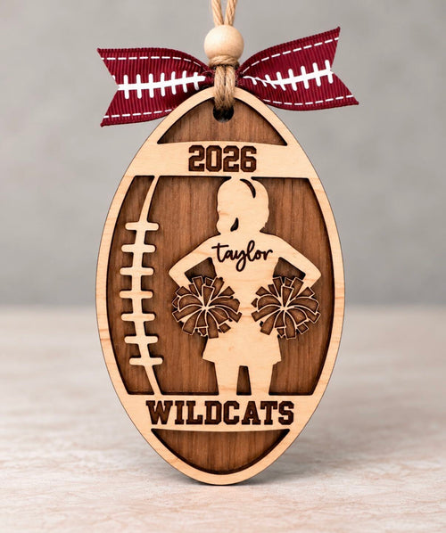 Wooden football shaped ornament with a cheerleader silhouette, her first name, team name, and year, tied with a football ribbon