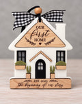 White house ornament with a wood roof engraved Our First Home, black windows and arched door, window boxes, and a wood base engraved Est. 2025 the beginning of our story, tied with a black and white gingham ribbon