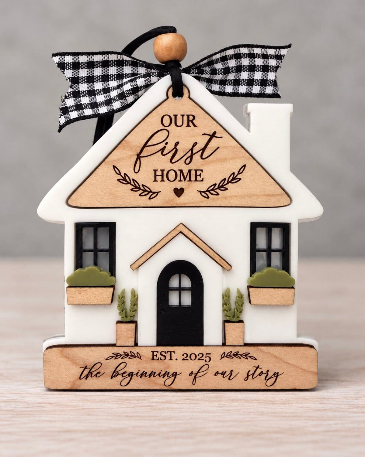 White house ornament with a wood roof engraved Our First Home, black windows and arched door, window boxes, and a wood base engraved Est. 2025 the beginning of our story, tied with a black and white gingham ribbon