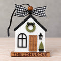 White farmhouse style house ornament with a black roof, green wreath, wood door, lantern, and potted tree on a wood base engraved The Johnsons, tied with a black and white gingham ribbon