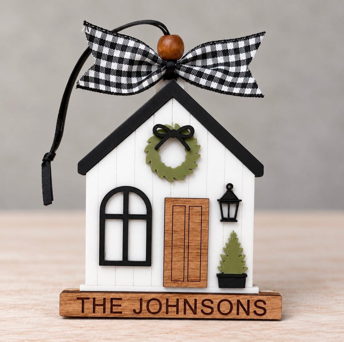 White farmhouse style house ornament with a black roof, green wreath, wood door, lantern, and potted tree on a wood base engraved The Johnsons, tied with a black and white gingham ribbon