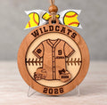Round layered wood softball ornament with a jersey engraved Jenna and number 9, a batting helmet, and a softball engraved 2026, with Wildcats and 2026 around the border, tied with a yellow softball ribbon