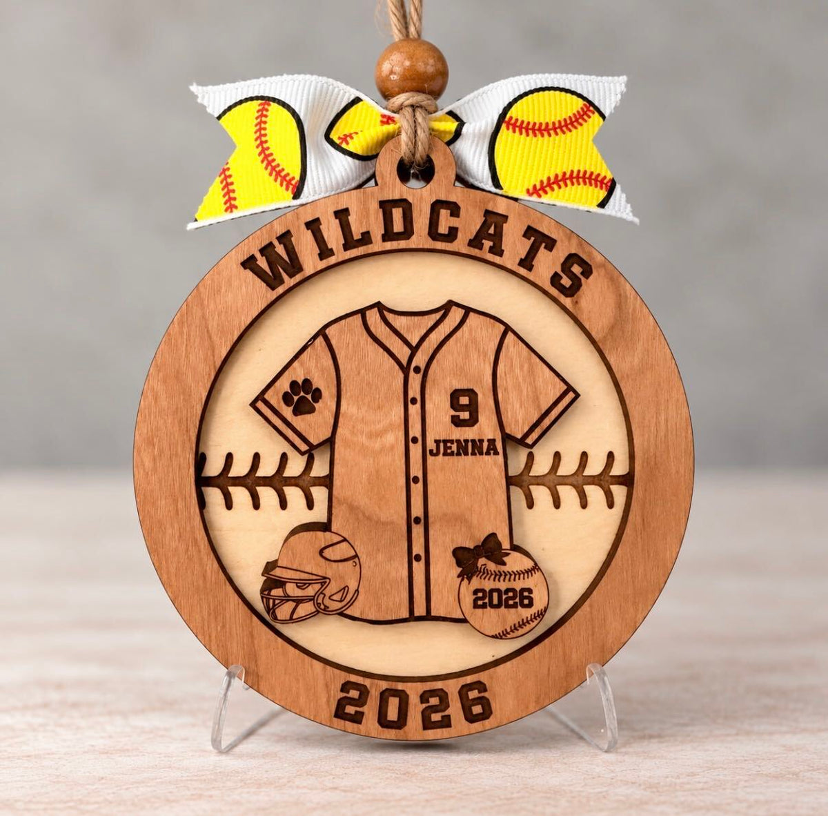 Round layered wood softball ornament with a jersey engraved Jenna and number 9, a batting helmet, and a softball engraved 2026, with Wildcats and 2026 around the border, tied with a yellow softball ribbon