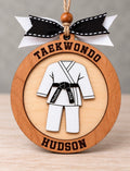 Round layered wood martial arts ornament with a white uniform and black belt engraved Hudson 2026, with Taekwondo across the top and Hudson across the bottom, tied with a black and white ribbon