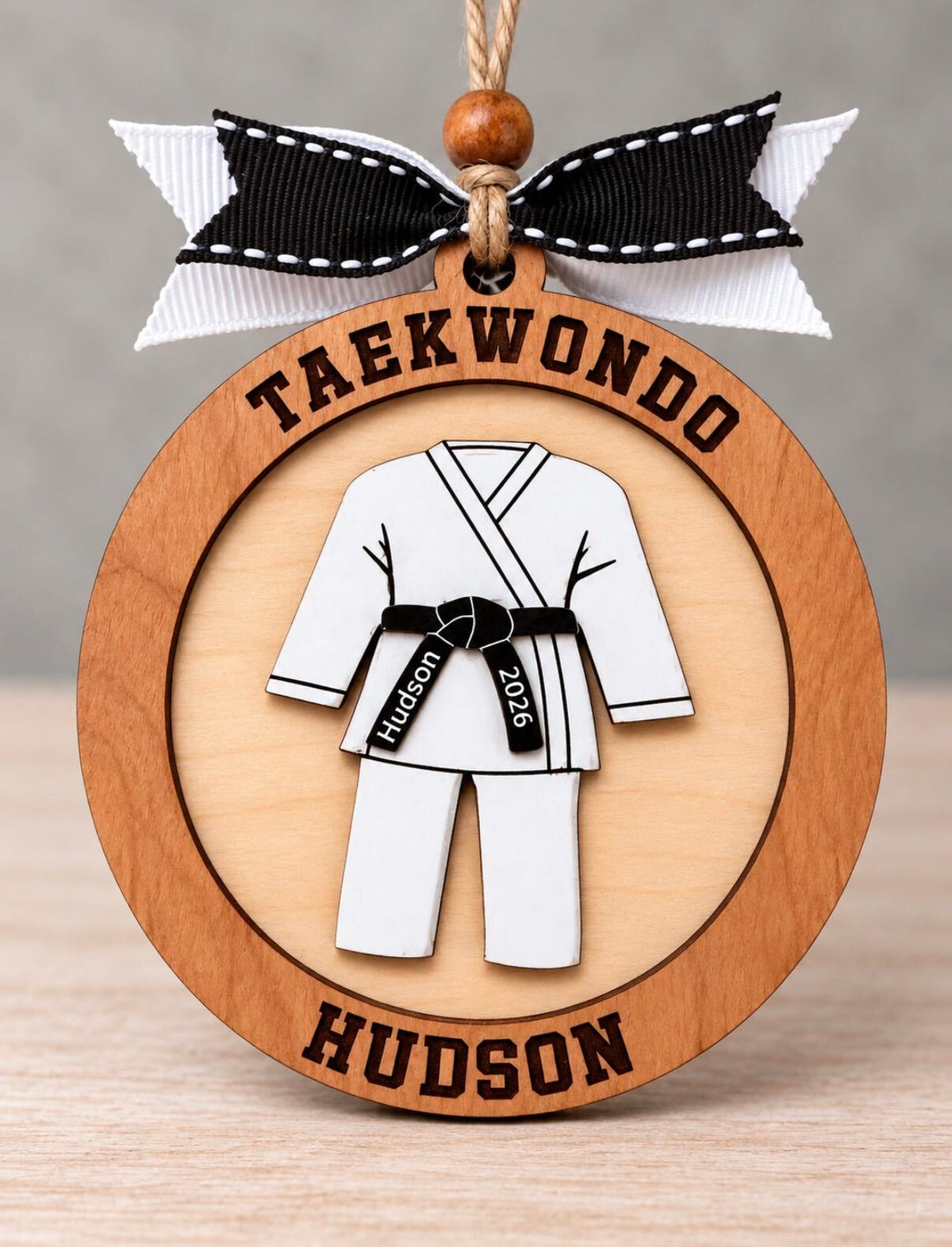 Round layered wood martial arts ornament with a white uniform and black belt engraved Hudson 2026, with Taekwondo across the top and Hudson across the bottom, tied with a black and white ribbon